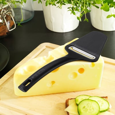 Cheese slicer on cheese, bread with slices, plants in bg. Alternatively: cheese slicer on wedge, bread with slices, potted plants.