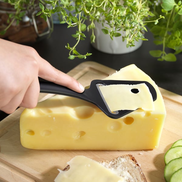 Person slicing cheese with black cheese slicer on wooden board. Fresh herbs in background.
