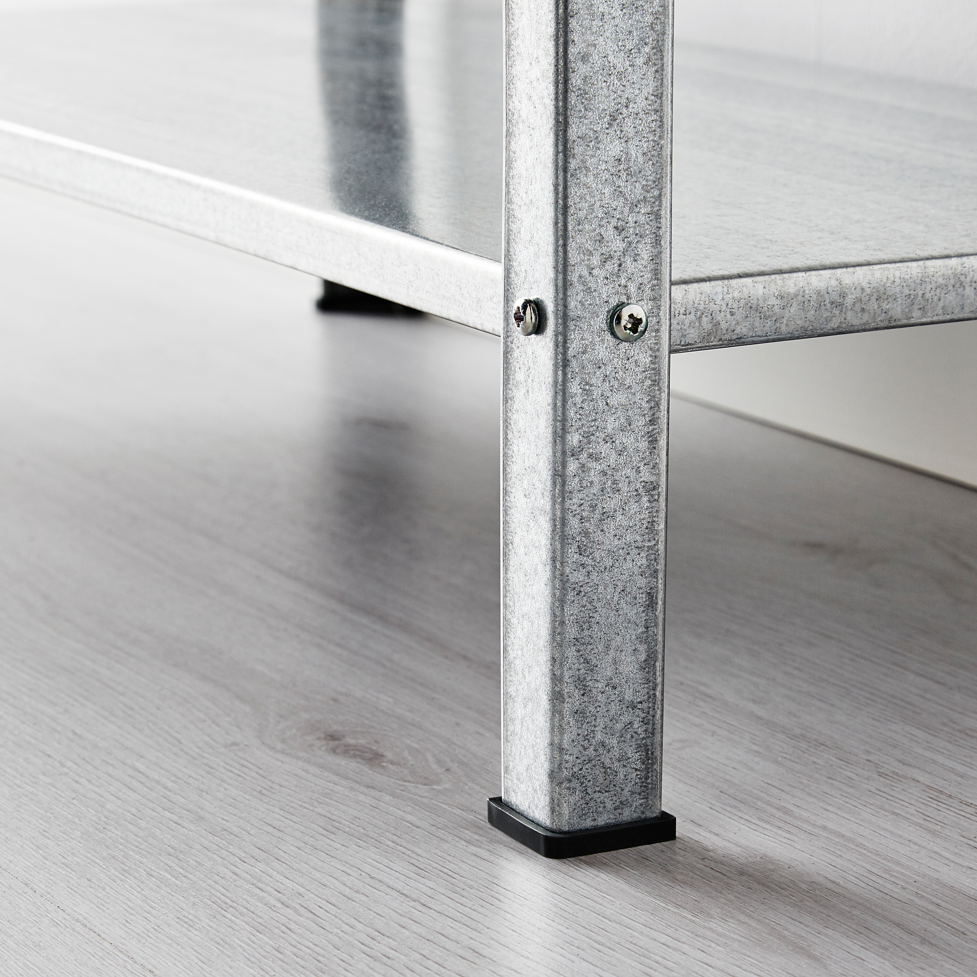 Image shows a close-up of a sturdy, galvanized metal shelf leg on a wooden floor. The leg adjustable black plastic feet for floor protection.