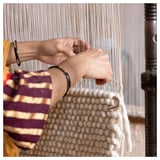 Person weaving a light-colored, handmade rug using a loom. Visible hands and part of the loom.