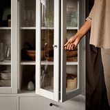 Person opening white glass-door cabinet, accessing woven basket, storing kitchen items.