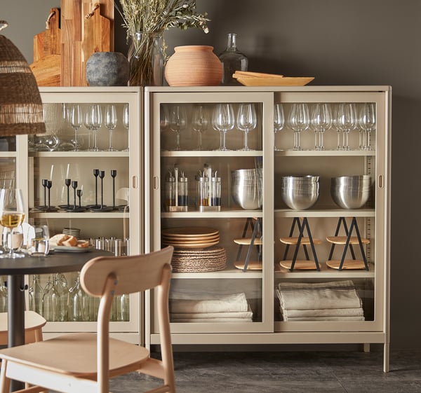 A modern glass cabinet, IDÅSEN, white, wine glasses, silverware, and bowls. Wooden chair near, decorative items on top.