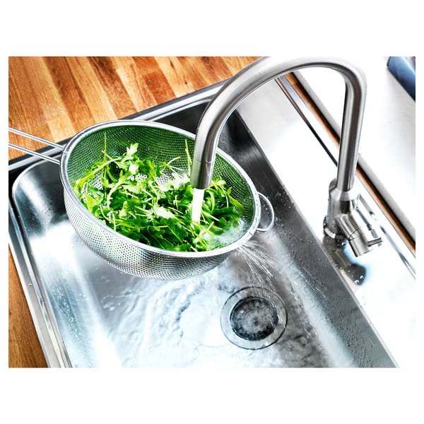 Stainless steel colander rinsing veg in sink. Faucet running, modern kitchen.