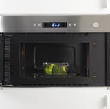 Stainless steel microwave with glass container of broccoli inside, showing its function of heating or defrosting food.