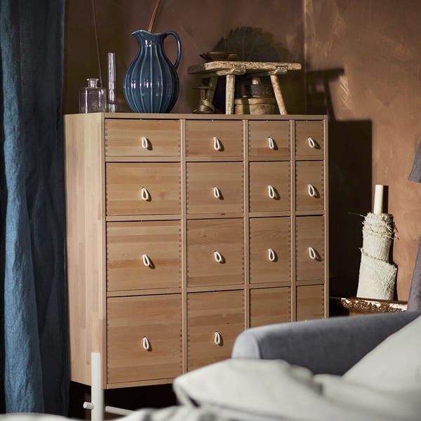 Wooden drawer unit, light brown with shell-shaped handles, holding blue vase, placed against warm-colored wall.