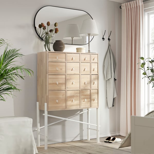 Modern light-colored wooden chest with white metal legs, featuring multiple small drawers, topped with a lamp and decorative items.