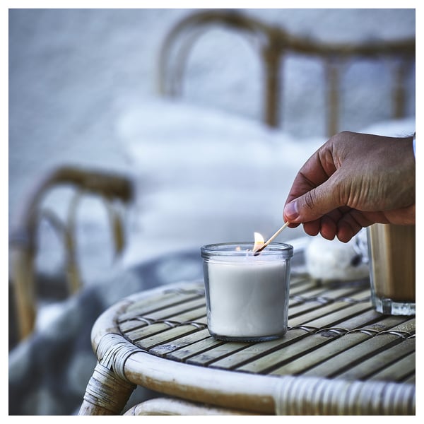 A hand lights a white JÄMLIK candle in a glass holder on a wicker table, creating a cozy atmosphere.