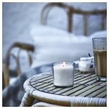 A small lit candle in a glass jar sits on a wicker table next to a glass of drink and a mug.