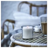 A burning white candle in a clear glass jar sits on a wicker table outdoors, beside a glass of frothy coffee.