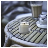 A small lit vanilla scented candle in a glass jar sits on a woven bamboo surface alongside a glass jar with dried flowers.