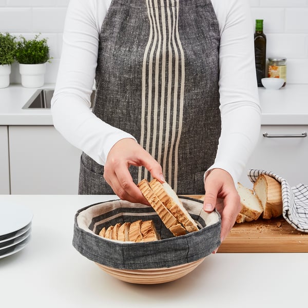 A person slices fresh bread into a woven proofing basket on a kitchen counter. The basket is used to shape and pattern bread during rising.