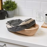 Wooden bread proofing basket with striped cloth on kitchen counter, near measuring cup and plant.