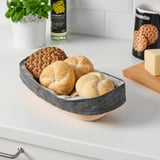 A grey bread proofing basket lined with cloth holds bread and crackers on a white kitchen countertop.