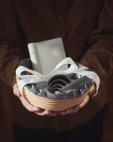 Hands hold a white JÄSNING basket with a silver ribbon, containing a white gadget and black kitchen tools.