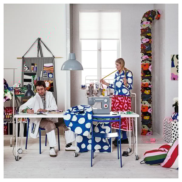 Two women crafting in bright, colorful studio; one sews blue polka-dot fabric, the other sketches at white table; large window, vibrant decorations.