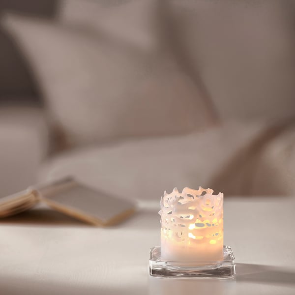 Small JUBLA white candle in glass, intricate lace design, burning on table.