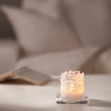 Small JUBLA white candle in glass, intricate lace design, burning on table.