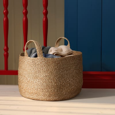 KÄLKEN jute basket with folded clothes, natural colour, round shape, sturdy handles, placed on wooden floor beside red chair.