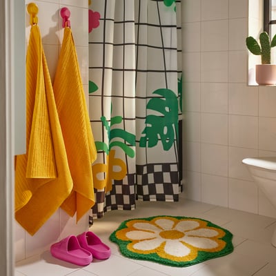 Bright bathroom with yellow towels, pink slippers, and a green/yellow flower-shaped KÄRRKNIPPROT mat. Decorative shower curtain with floral patterns.