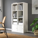 White KALLAX bookshelf with books, decor, and storage near gray wall. Wicker chair and plant nearby.