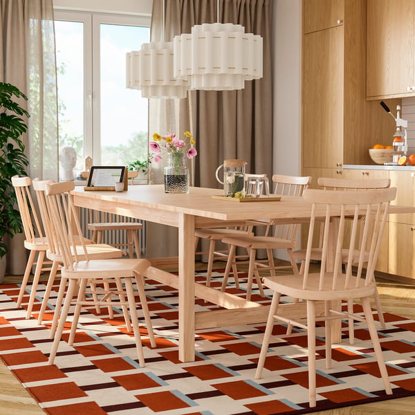 Modern dining room with large wooden table, 8 chairs, vibrant rug, stylish pendant lamp, bright decor, warm lighting, and large window.