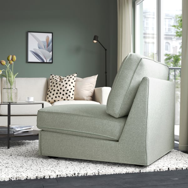 Modern living room with green walls. Gray sofa-bed is positioned near a large window. White coffee table with books and flowers is next to it.