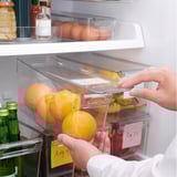 Person organizes oranges in clear fridge storage box with lid. Box has built-in handle and date note. Fridge well-organized with various items.