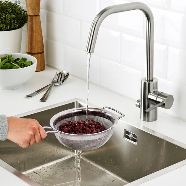 A hand holds a strainer under a kitchen faucet, rinsing red beans. A bowl of greens and utensils are on the counter.