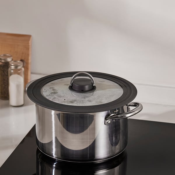 A stainless steel pot with a glass lid sits on a stove. The lid has a steam vent and a foldable handle, designed to fit various pot sizes.