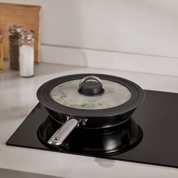 A black pot with a glass lid is on a stove, cooking food. The lid has a steam vent and a foldable handle.