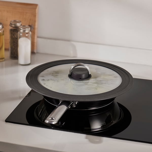 Black pot with glass lid on stove, steam vent visible, handle folded down.