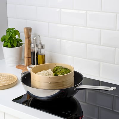 Black induction cooktop with bamboo steamer cooking broccoli and dumplings. White tiled backsplash.