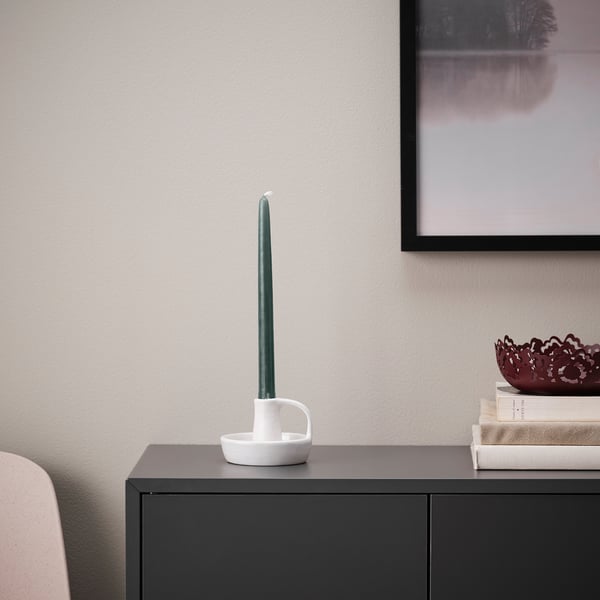 A tall green KLOKHET candle in white holder atop a dark cabinet, near a picture and stack of books, adding a cozy glow.