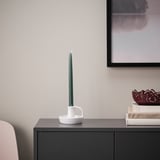 A tall green KLOKHET candle in white holder atop a dark cabinet, near a picture and stack of books, adding a cozy glow.