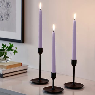 Three black candle holders with tall purple candles and warm light, near a plant and books.