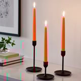 Three orange KLOKHET candles in black holders on table, lit, taller than nearby books and greenery.