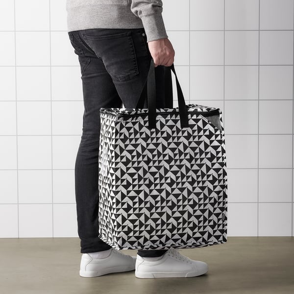 Person holding large KNALLA shopping bag with geometric pattern. The bag has handles and appears sturdy for groceries or laundry.