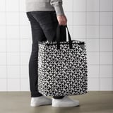 Person holding large KNALLA shopping bag with geometric pattern. The bag has handles and appears sturdy for groceries or laundry.