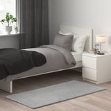 Small grey KNARDRUP rug, soft and sustainable, fits near clean white bed, contrasts dark blanket, adds warmth.