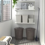 Two large grey KNODD storage bins with lids on floor, next to chair and shelves, with a blue broom.