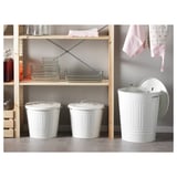 Three white bins with handles and lids, organized in front of a wooden shelf with glass containers and towels.