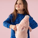 A young girl, smiling, hugs a soft, pink plush pig toy against a pink background. The person wears a blue sweater with dark spots.