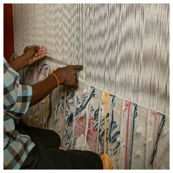 Person weaving KNYLHAVRE rug with colorful geometric patterns and fringes on loom. Hands manipulate threads, creating dynamic design.
