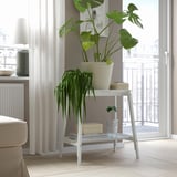 Plant stand near window with large, leafy plant & small blue-vased plant, book, beige box.