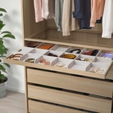 Wooden wardrobe drawer with gray organizer trays storing belts, ties, and jewelry.