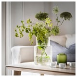 A clear glass vase holds green flowers, placed on a wooden table with small green candles nearby.