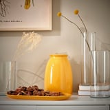 Yellow ceramic dish on white counter with decor, vases, and books. Botanical art in background.