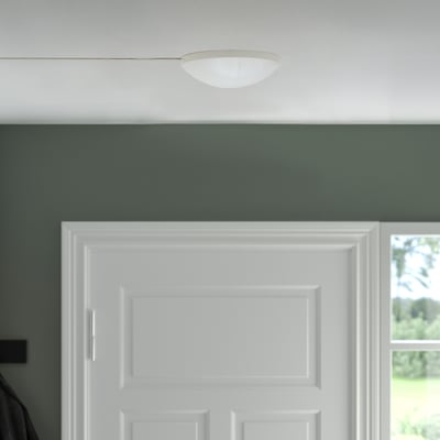 A KOPPLA white ceiling lamp plug in a green room. Its small, round, and neatly placed against the ceiling next to a white door.