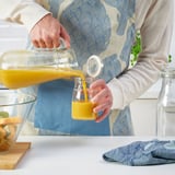Person pouring orange juice from KORKEN pitcher into square bottle with lid on countertop, fruits in background.