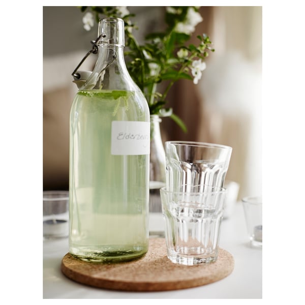 A glass bottle with a swing top stopper labeled elderberry on a coaster, alongside stacked clear glass cups.